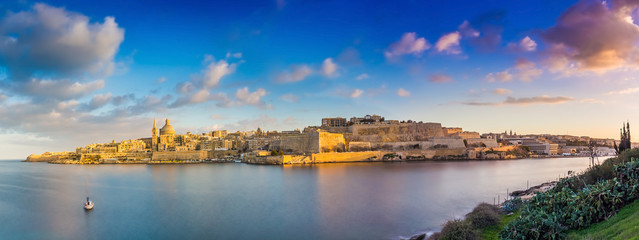 Fototapeta na wymiar Valletta, Malta - Panoramic skyline view of the ancient city of Valletta and Sliema at sunrise shot from Manoel island at spring time with sailing boat, blue sky, beautiful clouds and green grass