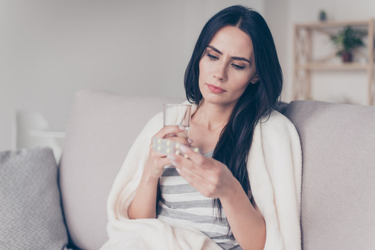 Portrait of troubled ill young woman sitting at home holding glass of water and reading discription of medication in her hand