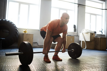 Middle-aged sporty man preparing to make exercise for biceps with help of barbell while having workout in modern gym