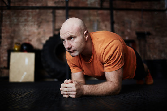 Bald middle-aged man doing plank exercise while having intensive workout in modern gym, full-length portrait