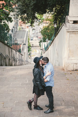 Handsome man kissing his brunette girlfriend in hat. Young modern hipster couple in love. Deep moment of a love story with a vintage look, city view in the background. 