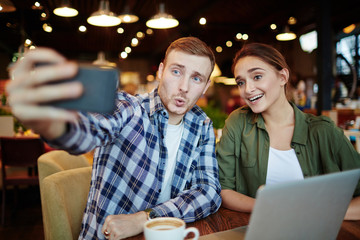 Having good time in lovely coffeehouse: handsome young man showing grimace while taking selfie with his smiling pretty friend, waist-up portrait