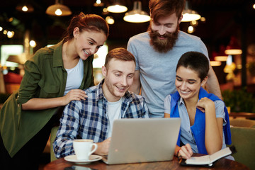 Four young friends hanging out in cozy small cafe: they enjoying delicious coffee and watching their favorite show on laptop