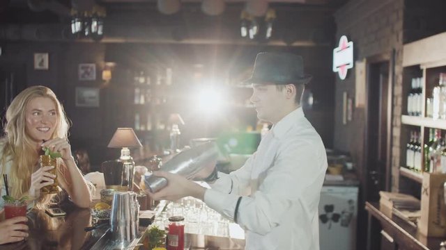 company of 20s friends are dring cocktails while bartender in hat is preparing drinks behind at the night party bar