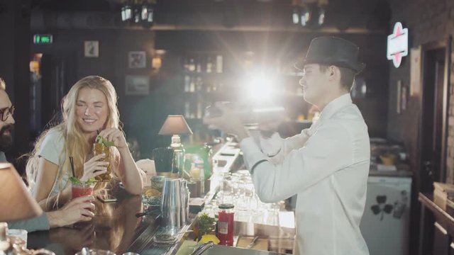 company of 20s friends are dring cocktails while bartender in hat is preparing drinks behind at the night party bar