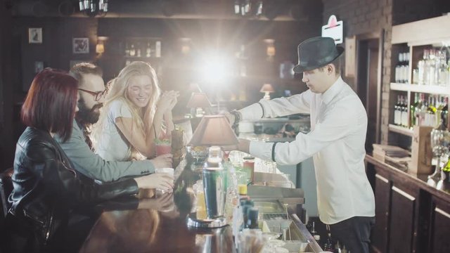 company of 20s friends are dring cocktails while bartender in hat is preparing drinks behind at the night party bar