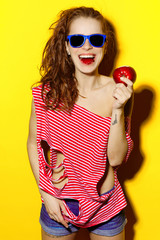 Beautiful young sexy girl in blue sunglasses and red striped T-shirt laughing and having fun and eating an apple on a yellow background