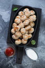 Black wooden serving board with roasted pelmeni in a frying pan, grey stone background, top view