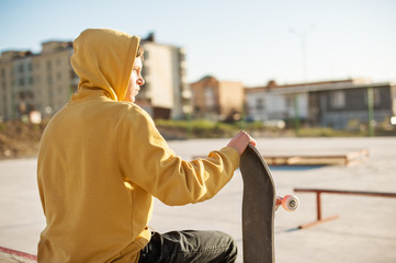 Close-up of a teenager dressed in a jeans hoodie sitting in a skate park and holding a skateboard