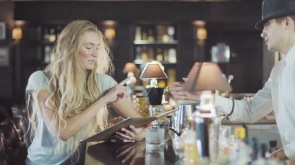 Bartender helps to blonde 20s woman make a choice in menu of bar