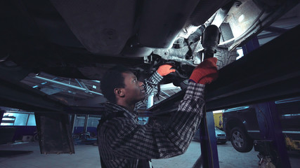 Fototapeta na wymiar African mechanic working on the underside of a car elevated on a hoist shining a bright light onto the chassis