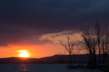 Sunset on a lake, with sun very low, dark and menacing clouds above, and some trees silhouettes in the foreground