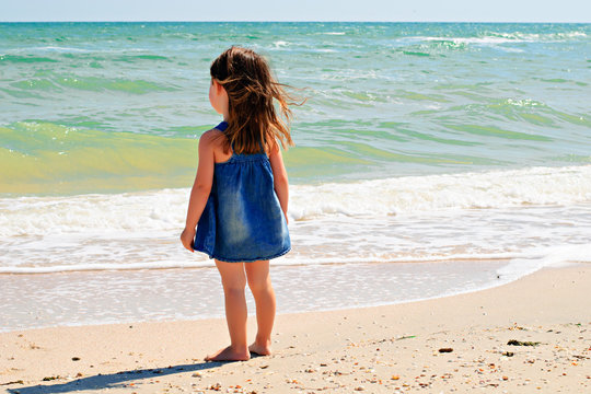 Cute little girl in denim dress standing alone on the beach looking at the sea