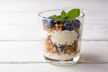 Granola with yogurt and blueberries in glass on white wooden table.