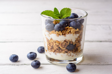 Granola with yogurt and blueberries in glass on white wooden table.
