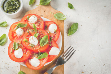 Italian food. Spring dinner for one person. Salad caprese - tomatoes, mozzarella cheese, fresh and dried herbs, basil, pesto sauce and raw pasta for cooking. On white stone table. copy space