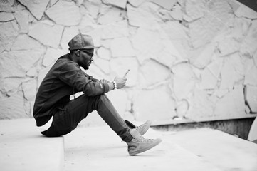Portrait of stylish african american man on sportswear, cap and glasses sitting on stairs with phone at hand. Black men model street fashion.