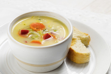 soup with healthy vegetables like carrot, red pepper and leek in a bright ceramic bowl with bread on a plate, white table, close up