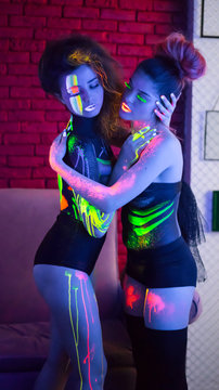 Two Sexy lesbian fashion modelshuggng each other. uv neon light with fluorescent glowing Body Art make-up . Low key dark image. Soft focus image.