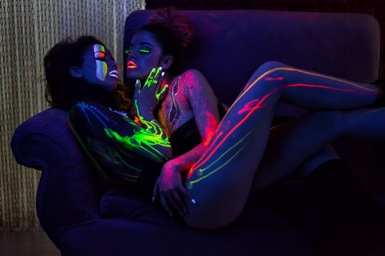 Sexy lesbian fashion models in uv neon light with fluorescent glowing Body Art make-up lying and kissing. Low key dark image. Soft focus image.