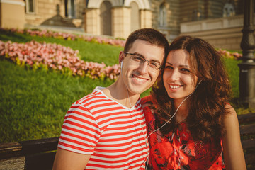 Obraz premium Young loving couple sitting on the bench in the center of the city, laughing, listening to music from smartphone