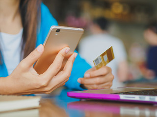 Pretty Young womans hands holding a credit card and using smartphone for online shopping. Online payment. Female working on laptop in a cafe.