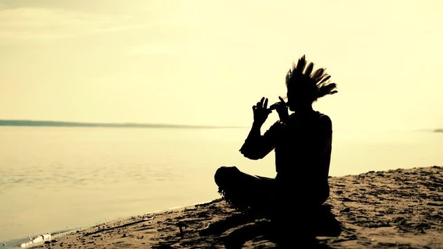 An Indian in a national costume plays a flute on the river bank 4k