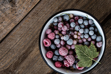 frozen berries, black currant, red currant, raspberry, blueberry in enamelled bowl decorated by currant leaf on wooden table in rustic style,  top view