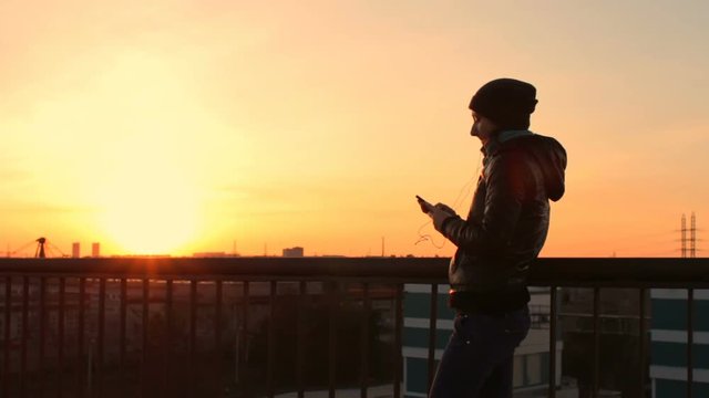 Young beautiful woman listening music with earphones standing on roof at sunset