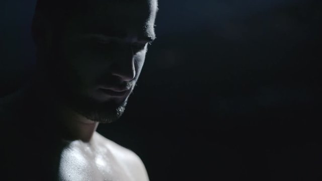 Close up of face of shirtless athlete standing in dark gym and contemplating 