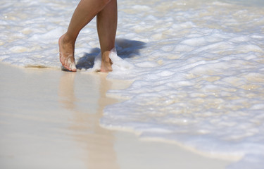 Vacation concept image. A caucasian woman is walking barefoot in the water line. Foam on white sand.
