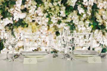 Glasses stand on table before wall of white flowers and greenery