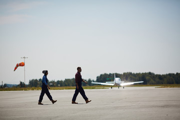 Wide shot image of two airport workers crossing runway field with plane flying up in background