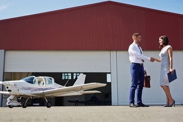 Portrait of two business people man and woman shaking hands standing by plane hangar in airport field