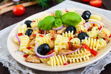 Pasta salad radiatori with chicken, black olives, blue onion, sweet pepper on a dark wooden background.