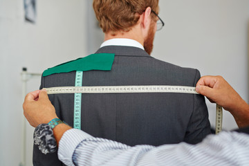 Old tailor taking back measurements from client in small atelier studio to make suit with jacket