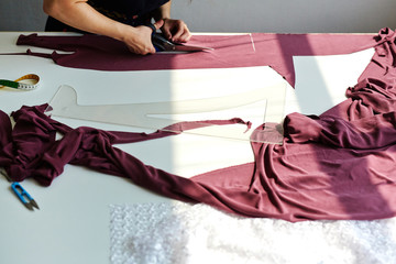 Background image of dressmakers studio: closeup of tailor cutting pink cloth to patterns on white workshop table with tools on top