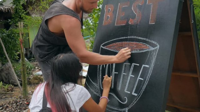 Young Hipset Couple Draws Art on Chalkboard Advertising Pillar near Coffee Shop. HD Slowmotion Closeup Footage.