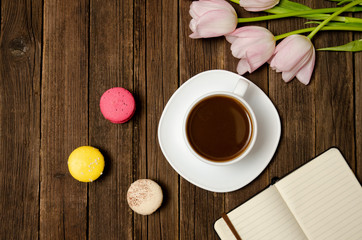 Fototapeta na wymiar Cup of coffee, macarons, pink tulips and notebook on wooden background. Top view