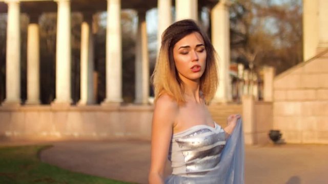 Beautiful model in silver and blue dress looks at camera, talks, smiles and poses during photo shooting outdoors with columns on background.