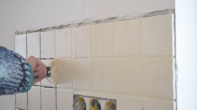 Hands of tile worker filling crevices between tiles with grout using soft rubber spattle