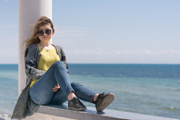 Blonde in jeans and sunglasses sits on a parapet near the sea shore