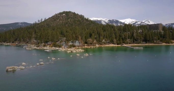 Flying over the surface of Lake Tahoe, flying towards the shore with private houses, mountains covered by snow on background