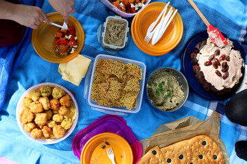 Unrecognizable people eating various picnic food: roasted vegetables salad, baba ghanoush, gluten-free crackers, foccacia bread, gluten-free and sugarfree dates cake. Top view. 
