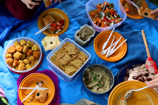 Unrecognizable people eating various picnic food: roasted vegetables salad, baba ghanoush, gluten-free crackers, foccacia bread, gluten-free and sugarfree dates cake. Top view. 
