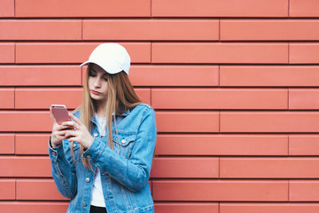 Portrait of stylish hipster girl in a cap and denim jacket street on a bright background. Girl on a background of red walls use their mobile phones