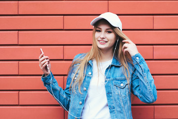Beautiful positive girl hipster holding a phone and listening to music on a bright red background. Smiling student wearing a denim jacket and cap-listening with headphones.
