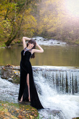 A brunette in a posh black dress posing by the river with a small waterfall.