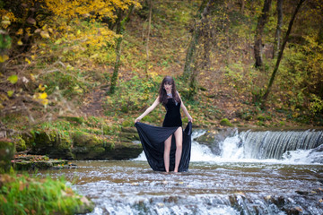 A brunette in a posh black dress posing by the river with a small waterfall.