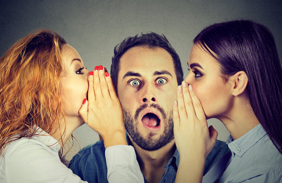 Two women telling whispering secret gossip in the ear to an amazed shocked man